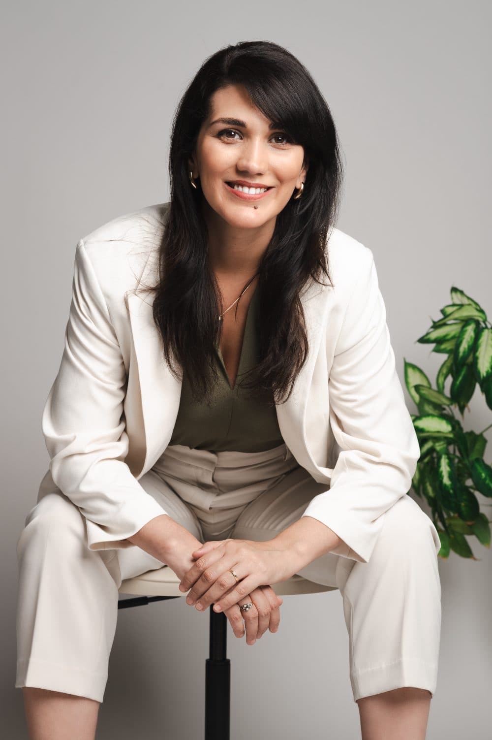 Isabel Fonseca in a white suit, seated on a stool with her hands clasped, smiling